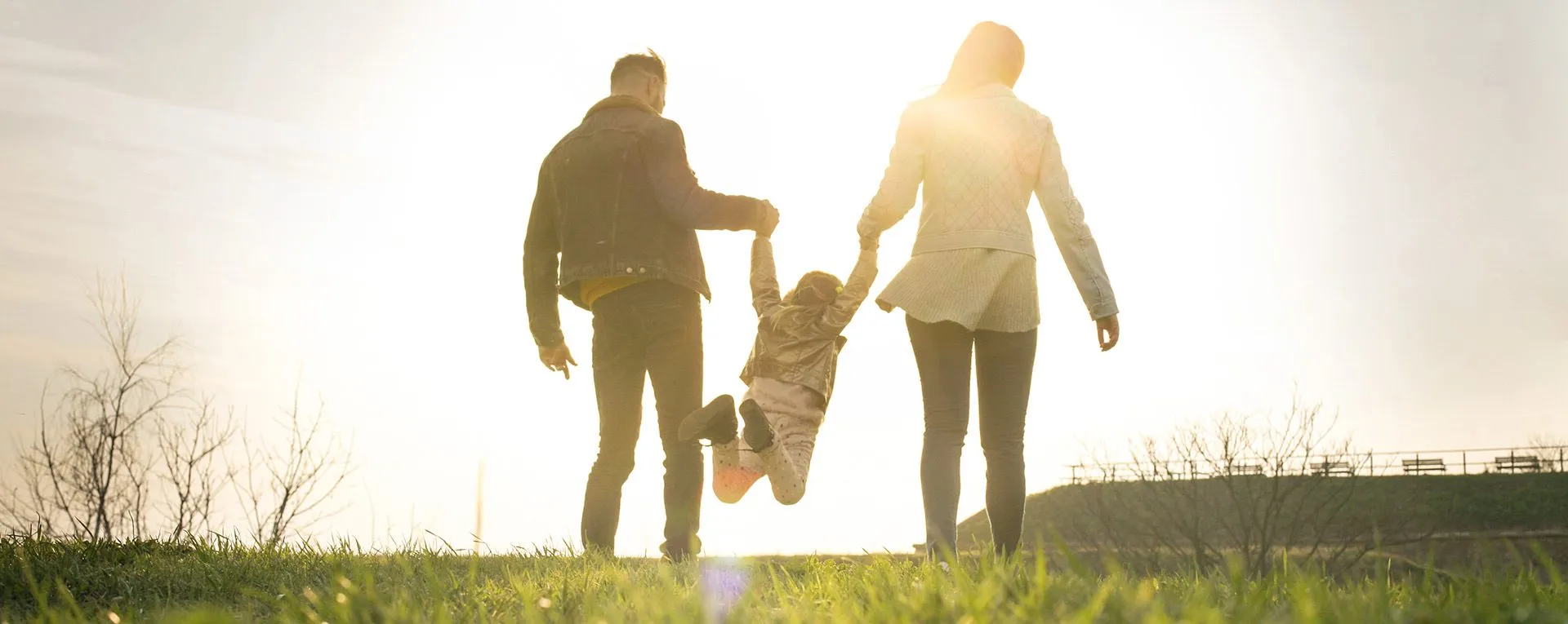 Two parents swinging their young child between them in a sunlit field